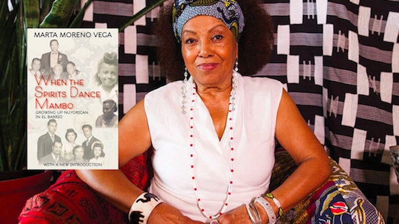 Dr. Marta Moreno Vega sits in a white shirt, bracelets and blue headscarf with hands together. Her book 
